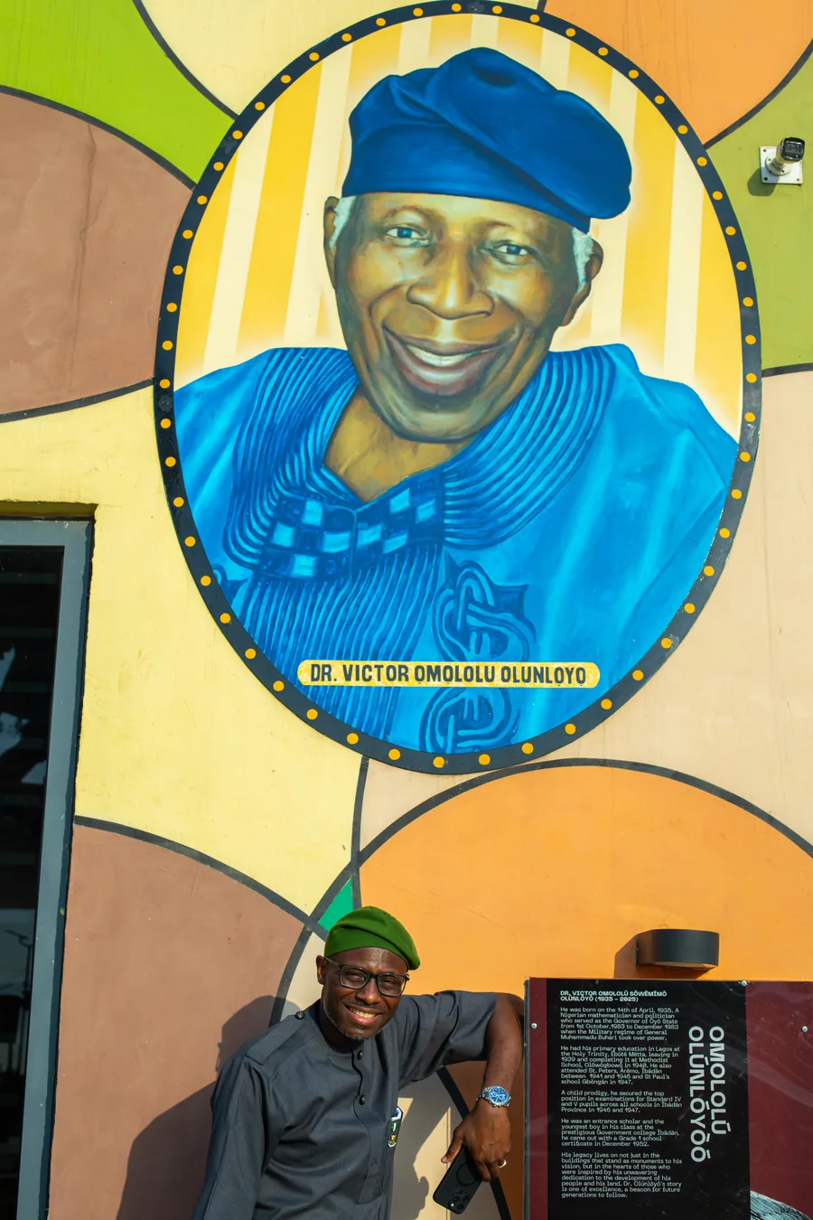 Ayobami standing before the mural of his father, Dr. Victor Omololu Olunlọyọ